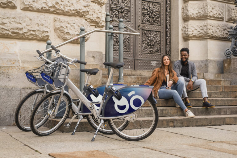 Next bike photo of a woman and a man sitting on steps waching 2 parked nexbikes -Micro Mobility